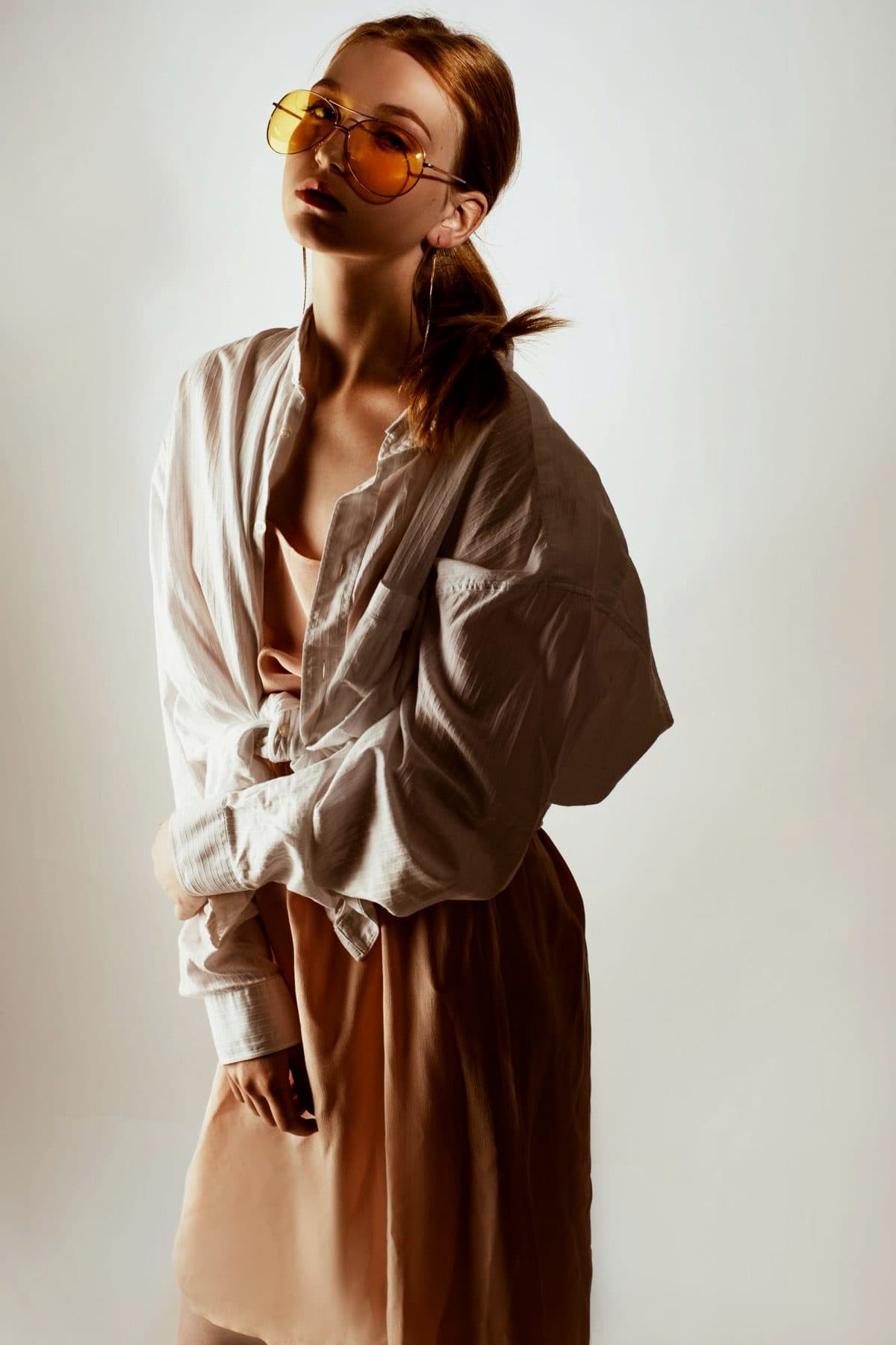 Oversized white shirt and amber glasses against a pale wall.